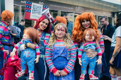 Comic-Con attendees hold Chucky dolls while dressed as Chucky in orange hair, striped colorful shirts, and overalls.