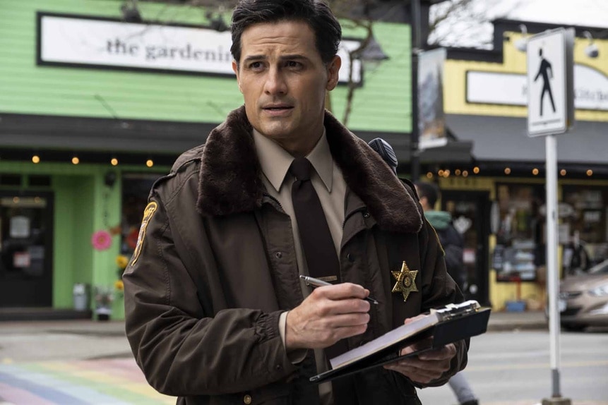 Joseph Rainier wears a police jacket while writing in a notebook in Resident Alien Episode 302.