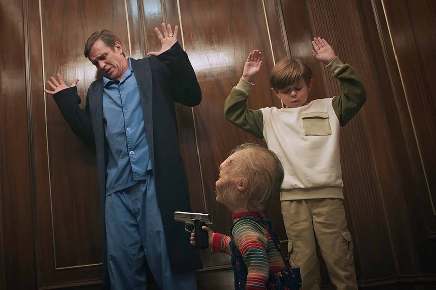 President James Collins (Devon Sawa) and Henry Collins (Callum Vinson) raise their arms as Chucky points a gun at them in Chucky Episode 306.