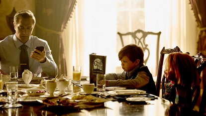 Grant Collins (Jackson Kelly) holds his phone as Henry Collins (Callum Vinson) reads a book next to Chucky at the dining table in Chucky 301.