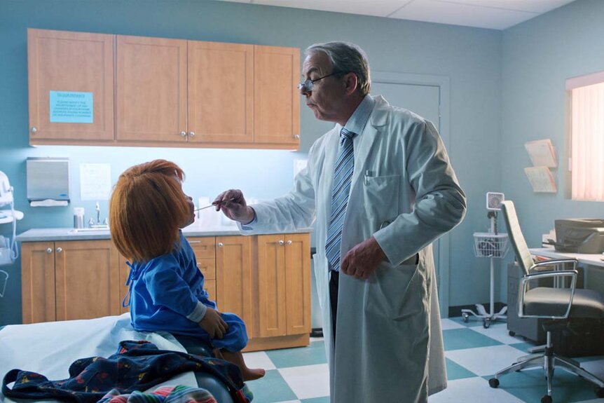 A doctor examines Chucky's mouth in an examination room in Chucky 303.