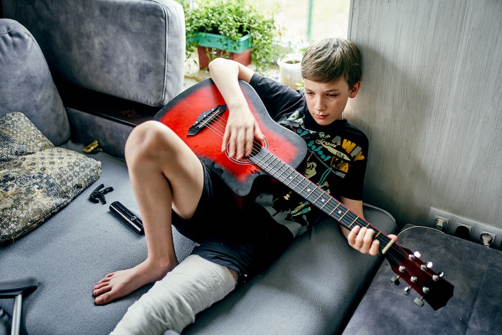 Boy with a broken leg playing a red guitar at home.