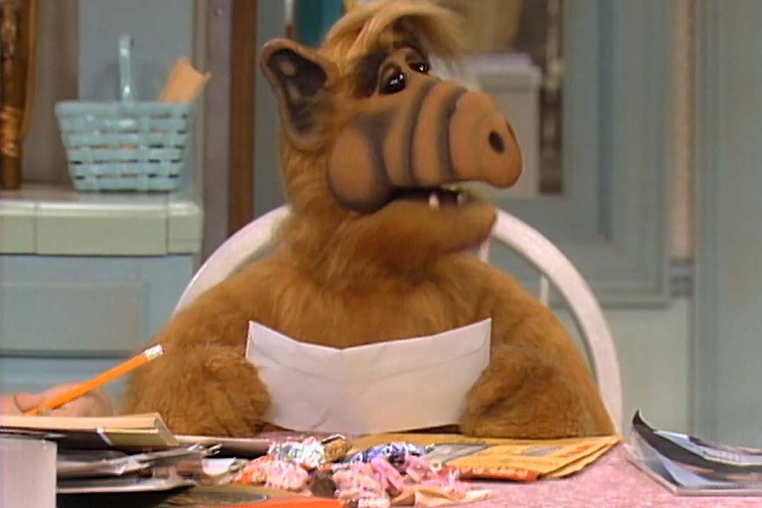 ALF sits at a table and holds a piece of paper.
