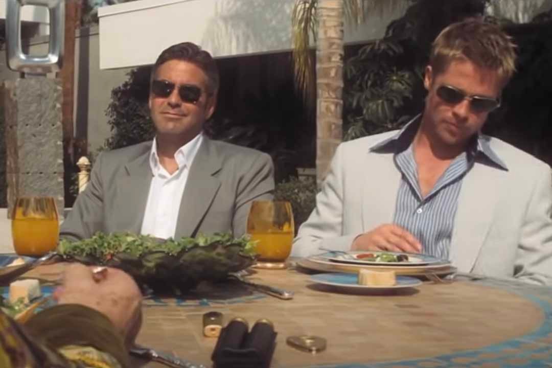 Danny (George Clooney) and Rusty (Brad Pitt) wear sunglasses and suits while sitting in front of food and drink in Oceans Eleven (2001).