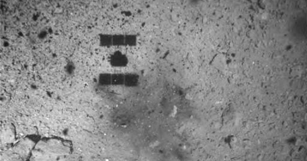 As Hayabusa2 ascends after (hopefully) retrieving a sample from the asteroid Ryugu, its shadow can be seen over the blast mark left from the projectile that dislodged surface rocks. Credit: JAXA