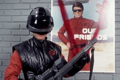 Visitor Soldier wearing a helmet and holding a gun in front of a vandalized poster from V: The Mini-Series