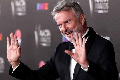 Sam Neill holds his hands up in a suit in front of a step and repeat.