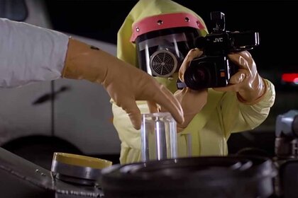 A hand holds a glass tube while a person in a protective suit points a camera at the hand in Back to the Future (1985).