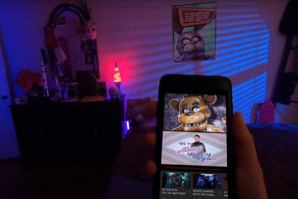 Freddy Fazbear appears on a video on a phone held by a hand in a dark room.