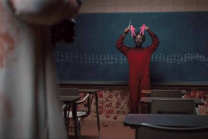 Red (Lupita Nyong'o) holds up red paper cutouts in front of a chalkboard with stick figures and red handprints under in Us (2019).