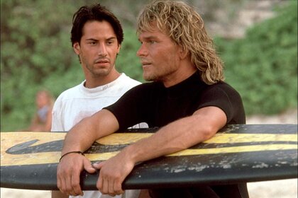 Johnny Utah (Keanu Reeves) watches Bodhi (Patrick Swayze) hold a surfboard in Point Break (1991).