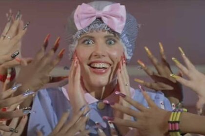 Valerie (Geena Davis) touches her face while wearing a bow bonnet as many hands with manicured fingers stick out toward her face in Earth Girls Are Easy (1988).