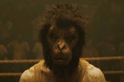 A monkey with the body of a man wears a yellow shirt in Monkey Man (2024).