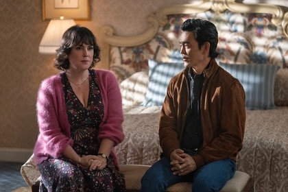 Melanie Lynskey and John Cho sit on a bench at the foot of a bed in Poker Face Season 2.