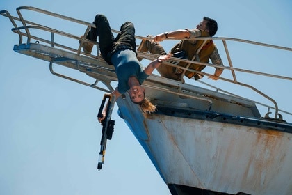 Zora Bennett (Scarlett Johansson) hangs off a ship with a large gun in her hand as Dr. Henry Loomis (Jonathan Bailey) holds onto her in Jurassic World Rebirth (2025).