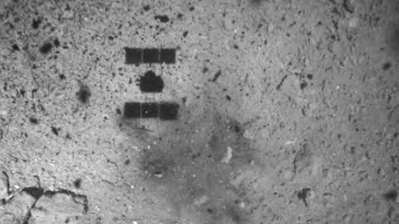 As Hayabusa2 ascends after (hopefully) retrieving a sample from the asteroid Ryugu, its shadow can be seen over the blast mark left from the projectile that dislodged surface rocks. Credit: JAXA
