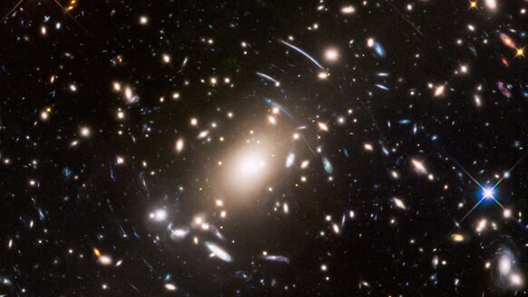 The galaxy cluster S1063 is a collection of hundreds of galaxies and is strewn with dark matter. This acts as a gravitational lens, distorting the light form more distant galaxies into arcs, which you can see throughout the center of this Hubble image. 