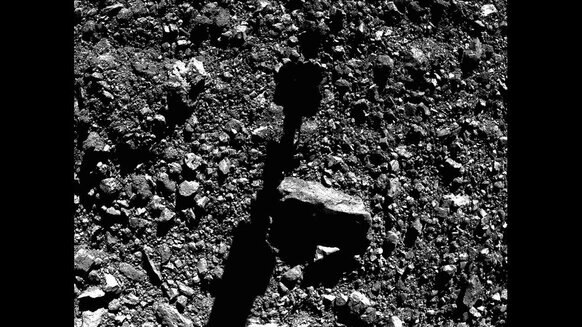 Moments before sample collection, the OSIRIS-REx boom and sample collector shadow stretched across the surface of the asteroid Bennu. Most of those rocks are a few centimeters across. Credit: NASA/Goddard/University of Arizona/Lockheed Martin