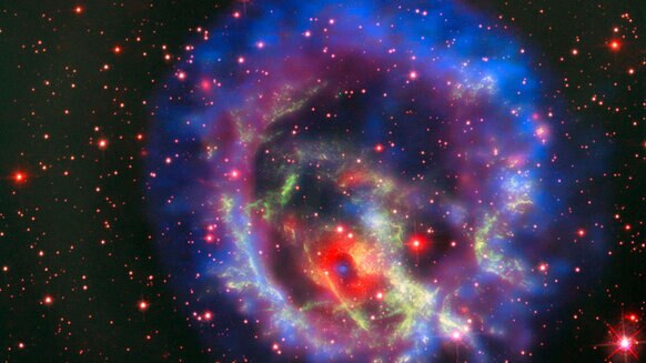 The supernova remnant 1E 0102.2-7219 seen by three telescopes: The red background (note the stars) from Hubble, the Very Large Telescope in green, and X-rays seen by Chandra X-ray Observatory in blue and purple.
