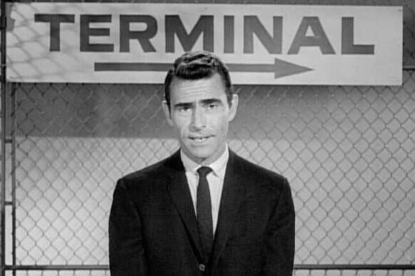 Rod Serling wears a suit and stands in front of sign that says "Terminal" on The Twilight Zone.