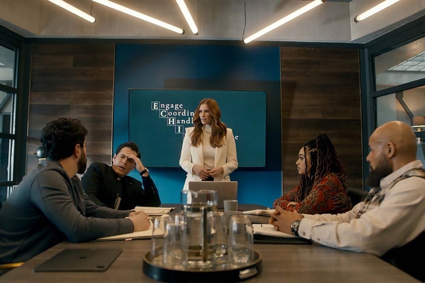 The cast of SurrealEstate sits at a conference table and watches Susan Ireland (Sarah Levy) stand at the head of the table in SurrealEstate 201.