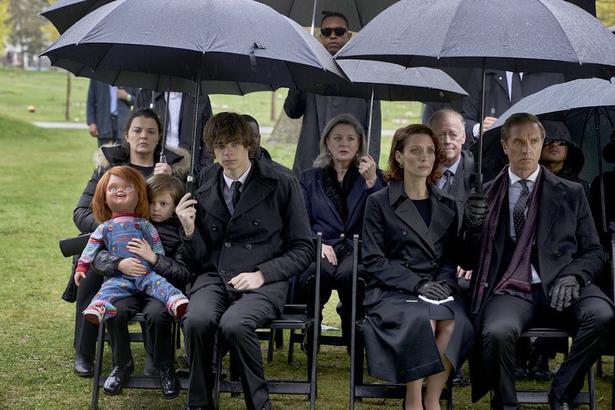 The cast of Chucky sits under a cover of black umbrellas at a funeral.