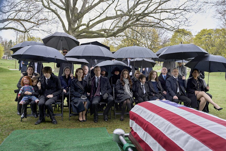 The cast of Chucky sits under a cover of black umbrellas and watches an American flag shrouded casket at a funeral.