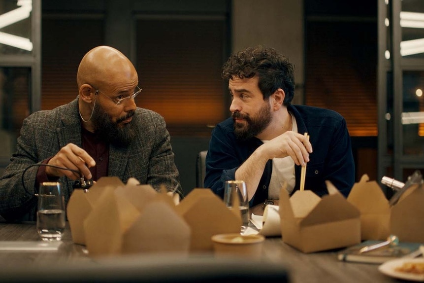 (l-r) August Ripley (Maurice Dean Wint) and Father Phil Orley (Adam Korson) speak to each other in front of a takeout feast on a table in SurrealEstate 203.