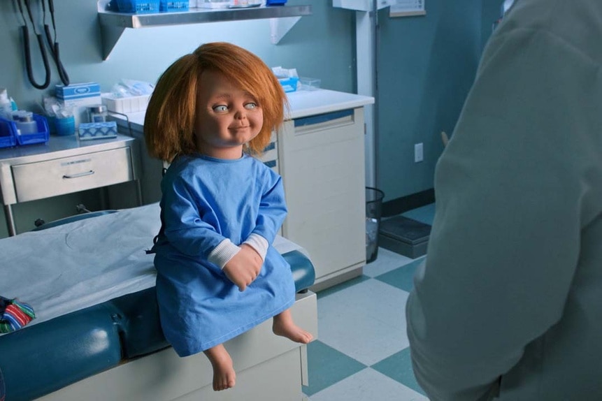 Chucky sits on an exam room table in a blue patient's gown and faces a doctor in Chucky 303.