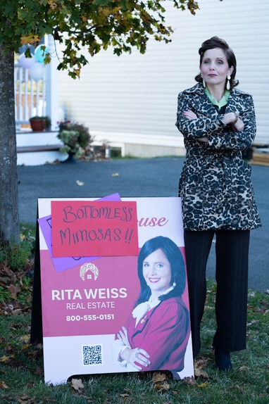 Rita Weiss (Alison Brook) stands near her own real estate advertisement sign in SurrealEstate 206.