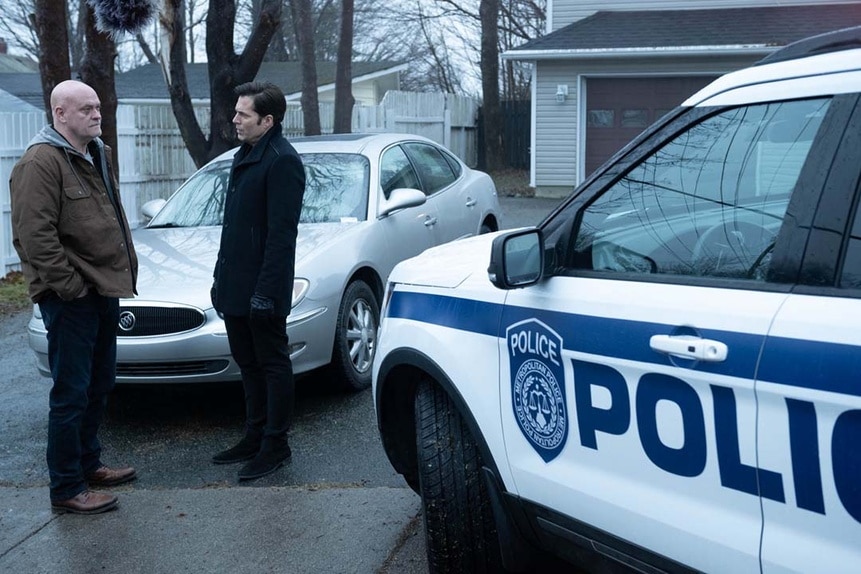 Morris Truby (James Collins) and Luke Roman (Tim Rozon) speak in a driveway next to a police car in SurrealEstate 207.