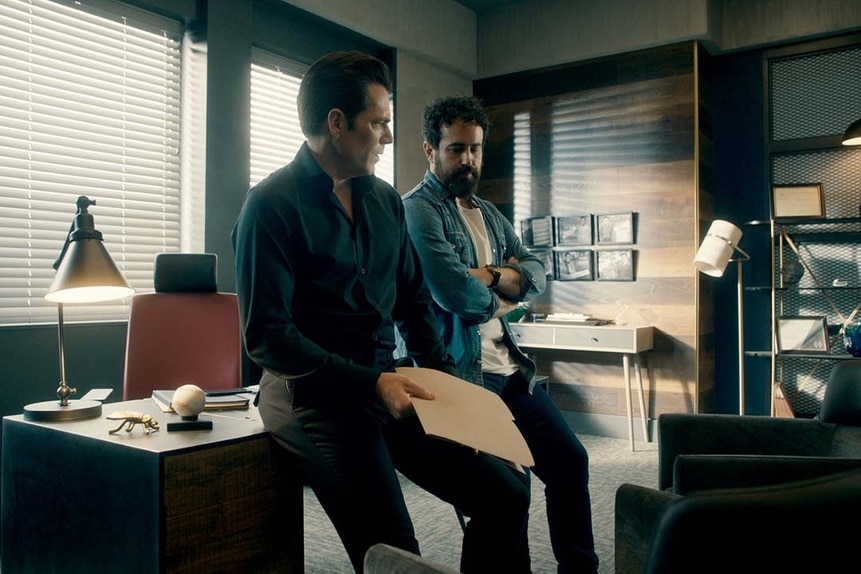 (l-r) Luke Roman (Tim Rozon) and Father Phil Orley (Adam Korson) sit on a desk together in an office in SurrealEstate 205.