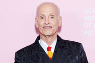 John Waters smiles in front of a pink backdrop.