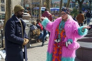 Flora (Melissa McCarthy) (R) wears a pink and teal coat while shrugging at Bernard (Paapa Essiedu) in Genie (2023).