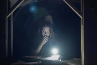 Anya (Holland Roden) reads with a flashlight in an attic while a monster stands behind her in Mother, May I? (2023).