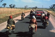 A gang of bikers follow a red car on the road in Mad Max (1979).