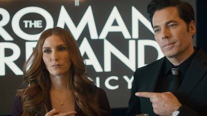 (l-r) Susan Ireland (Sarah Levy) and Luke Roman (Tim Rozon) point at each other in front of a "The Roman and Ireland Agency" sign in SurrealEstate 210