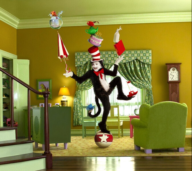 The Cat in the Hat (Mike Myers) balances on a ball in a green living room while balancing various objects like a fishbowl, cake, and umbrella in The Cat in the Hat (2003).