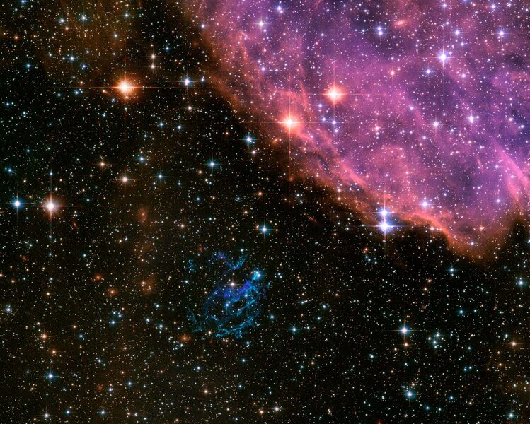 A wide field of view Hubble image of the supernova remnant 1E 0102.2-7219 (below center, blue) shows it lies just a few dozen light years from the huge star-forming nebula N76 in the Small Magellanic Cloud, a companion galaxy to the Milky Way. 
