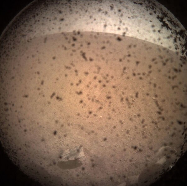 The very first image from NASA’s Mars InSight lander sitting on the surface of Mars. Dust particles on the dust cover obscure things somewhat, but the surface of Mars and the curved horizon (due to the wide-angle lens) are obvious, including a rock nearby