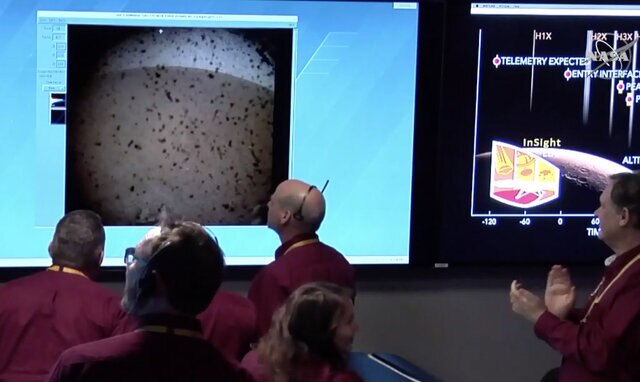 Screen grab from NASATV feed showing the Mars InSight ground team looking at the first image from Mars. The dust cover is on, so there is dust on it, but through that you can see the Martian horizon and small rocks. Credit: NASA