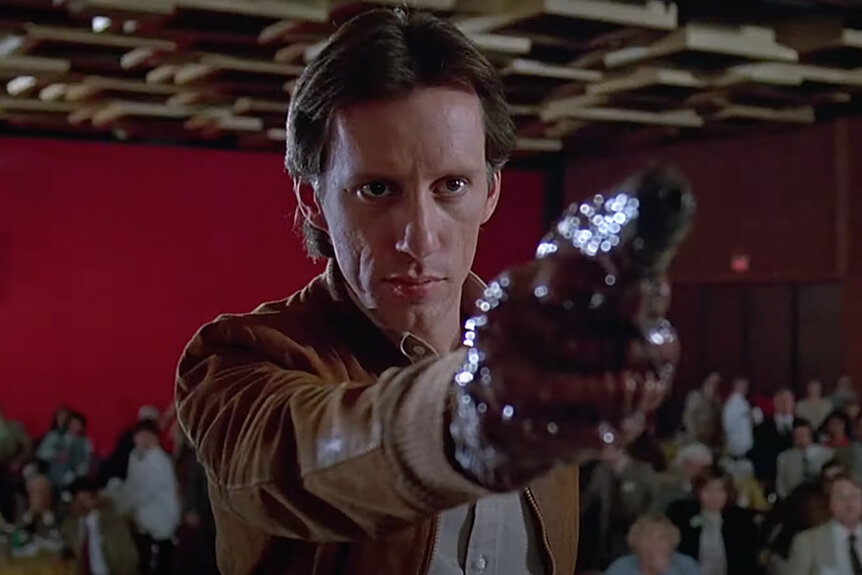 Max Renn (James Woods) holds up and points his flesh gun to someone menacingly in Videodrome (1983).