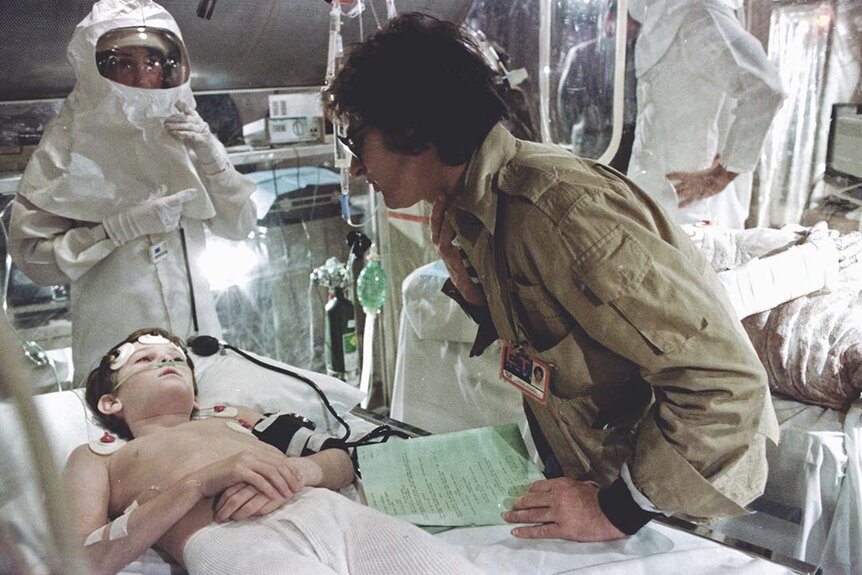 Steven Spielberg directs Henry Thomas on a hospital bed with hazmat suited person standing near during shooting of the emotional sequence in which E.T. appears to have died.