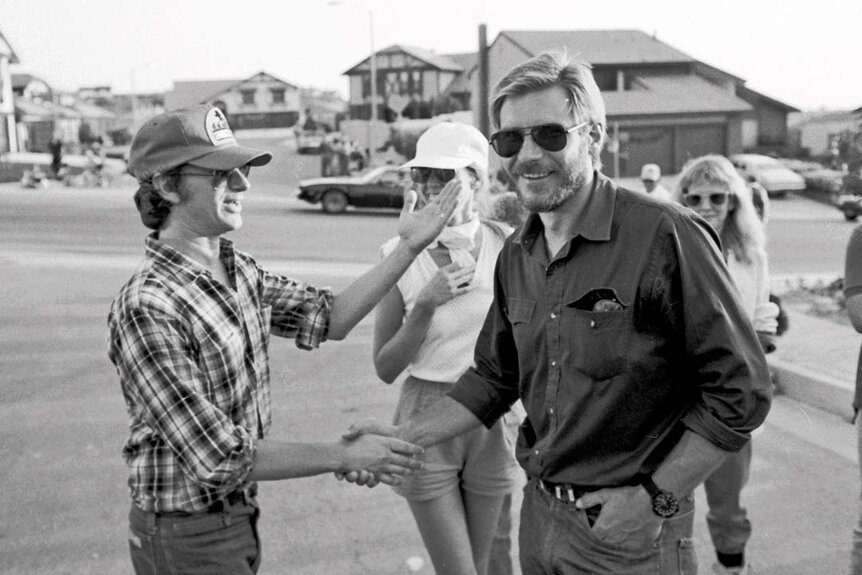 Steven Spielberg shakes hands with Harrison Ford as women watch.