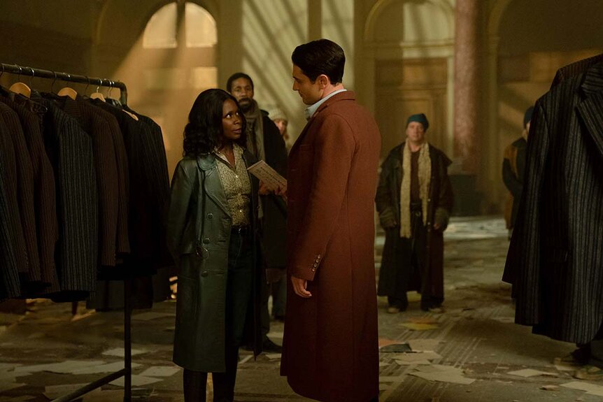 (l-r) Maizie (Zainab Jan) speaks to Winston (Colin Woodell) near a clothes rack in The Continental: From the World of John Wick Night 3 as other people watch on.