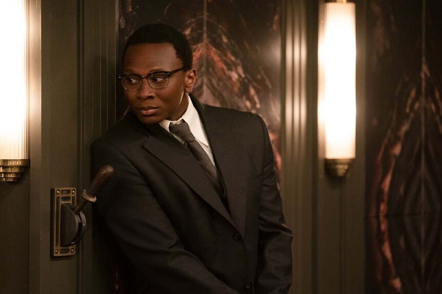 A suited Charon (Ayomide Adegun) leans on a wall near a lever in The Continental: From the World of John Wick Night 3.