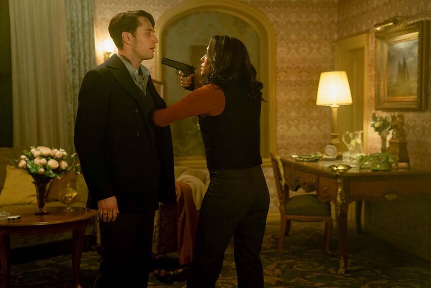 KD (Mishel Prada) points a gun at Winston's (Colin Woodell) face in an adorned hotel room in The Continental: From the World of John Wick Night 3.