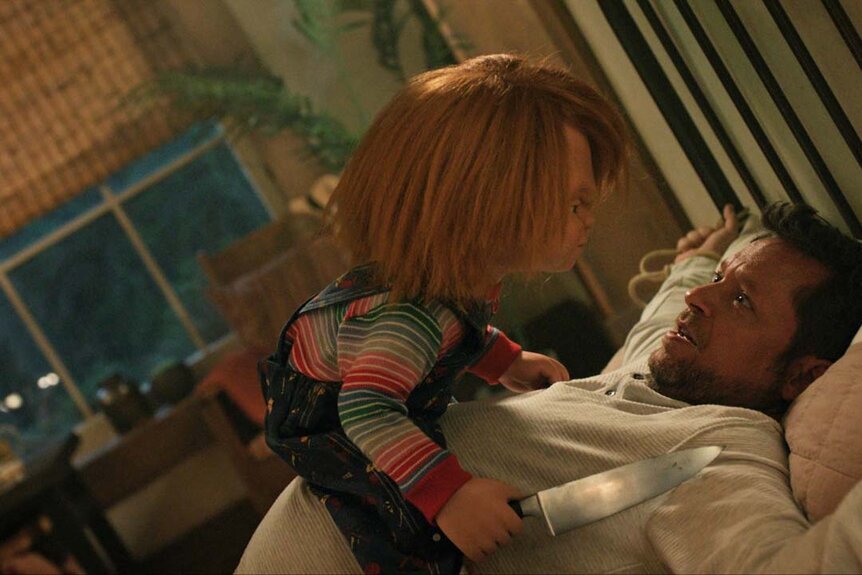 Chucky holds a knife while on top of a man in a bed in Chucky 303.