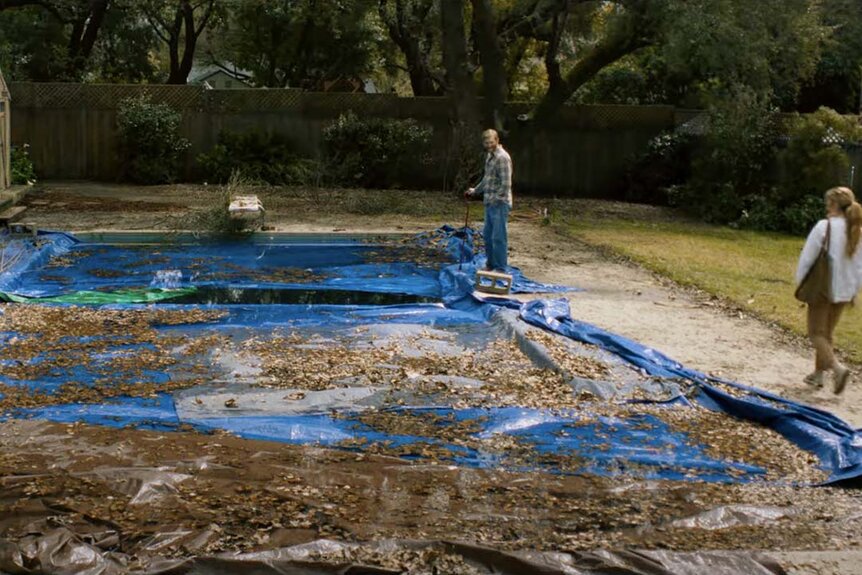 A man stands next to a pool covered in leaves and blue tarp as a woman walks toward him in Night Swim (2023).