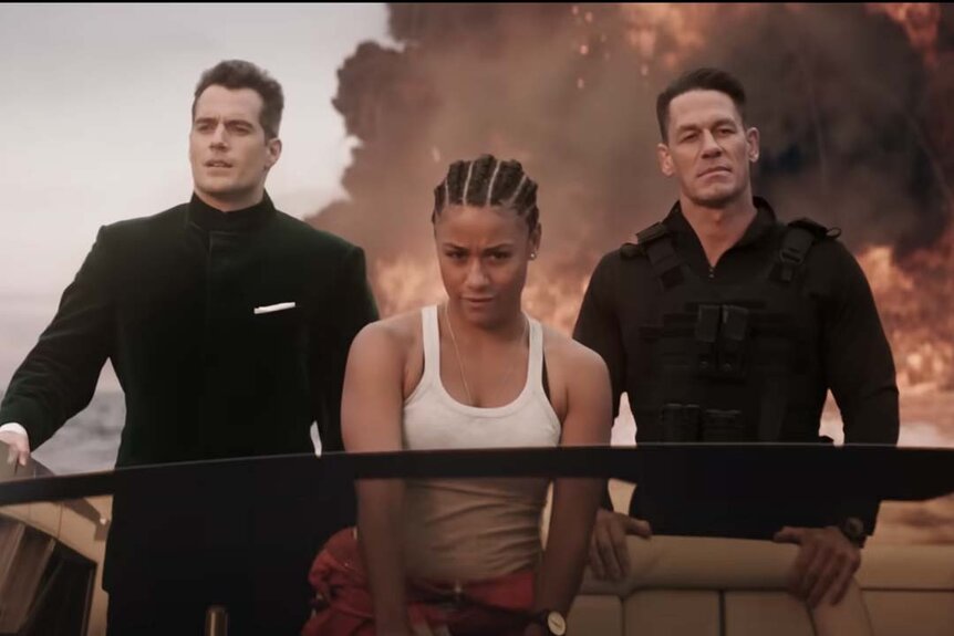 Henry Cavill, Ariana DeBose, and John Cena speed away on a boat while an explosion takes place in the background in Argylle (2024).